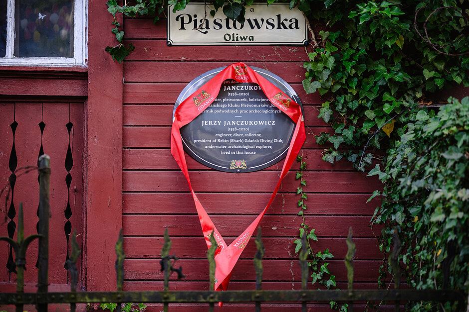 Na czerwonej, drewnianej elewacji budynku w Oliwie znajduje się okrągła tablica pamiątkowa poświęcona Jerzemu Janczukowiczowi (1938-2020), pionierowi nurkowania i badaczowi archeologii podwodnej. Tablica jest częściowo zasłonięta czerwoną wstęgą z herbami Gdańska, co wskazuje na moment tuż po jej odsłonięciu. Nad tablicą widoczna jest tabliczka z nazwą ulicy „Piastowska”, a prawą część ściany porasta gęsty bluszcz. W dolnej części kadru znajduje się ciemne, metalowe ogrodzenie.