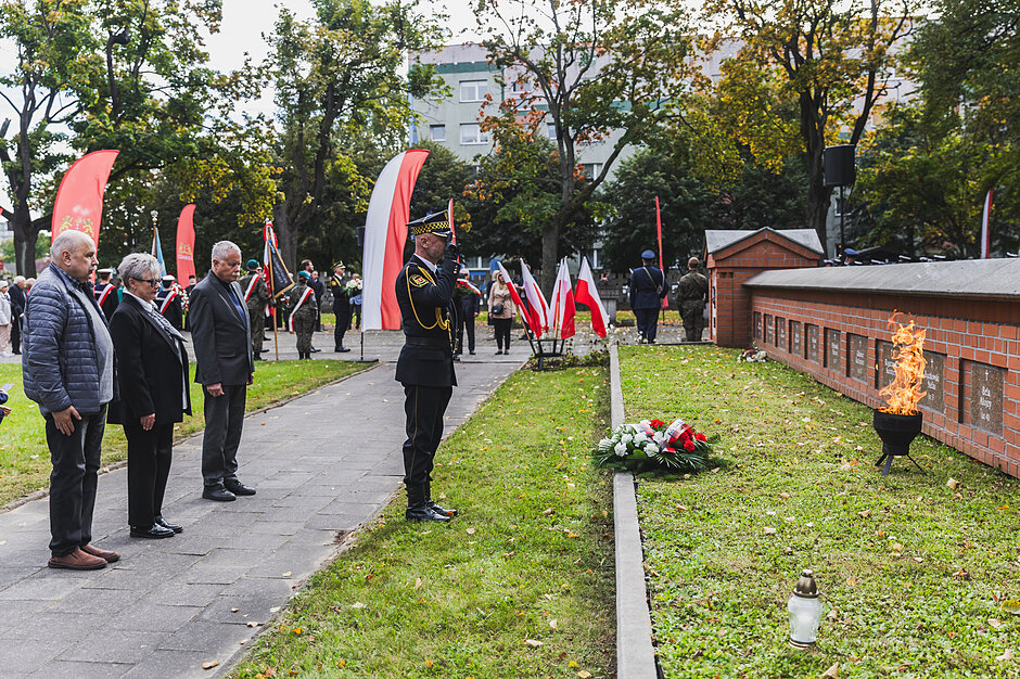trzy osoby stoją przed grobem, na którym leży wiązanka, przed nimi stoi salutujący strażnik miejski