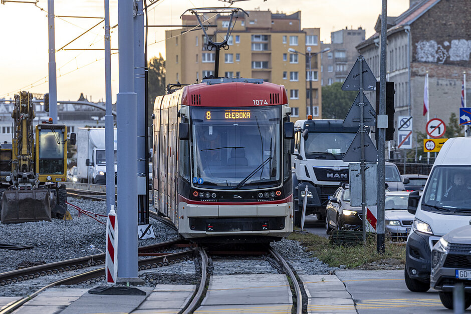 czerwono-biały tramwaj na torze tramwajowym w mieście. tramwaj ma numer 8 z przodu i napisane na nim słowo głęboka  wydaje się być nowoczesnym tramwajem elektrycznym o eleganckim wzornictwie. po prawej stronie jest ruchliwa ulica z samochodami i ciężarówkami.