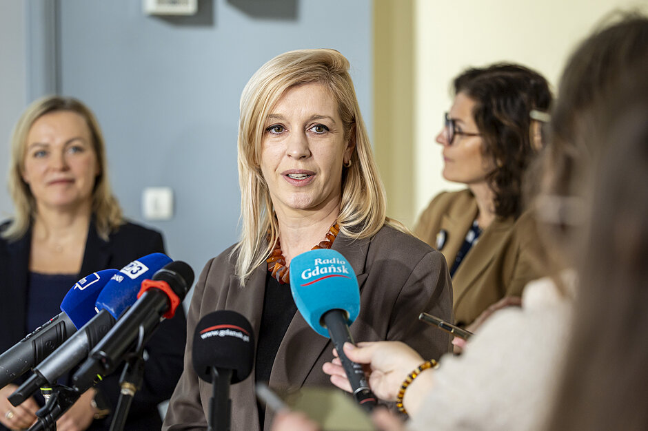 kobieta mówi do mikrofonu, za nią stoją dwie inne panie