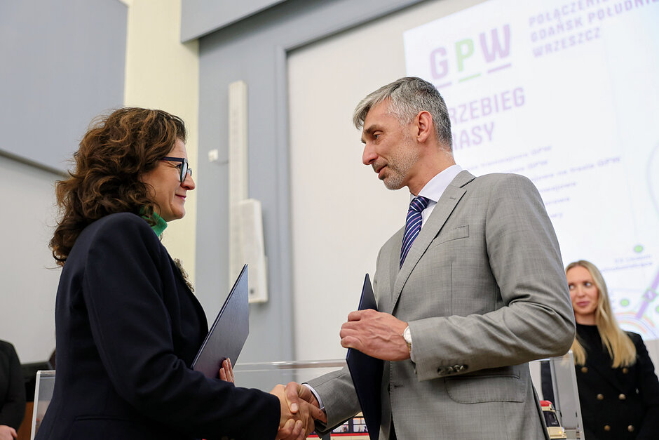 na zdjęciu kobieta i mężczyzna w średnim wieku, ściskają sobie dłonie, każde trzyma granatową teczkę w ręce