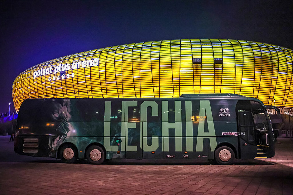 Autokar Lechii stoi na tle stadionu, rozjarzonego iluminacją na bursztynowy kolor