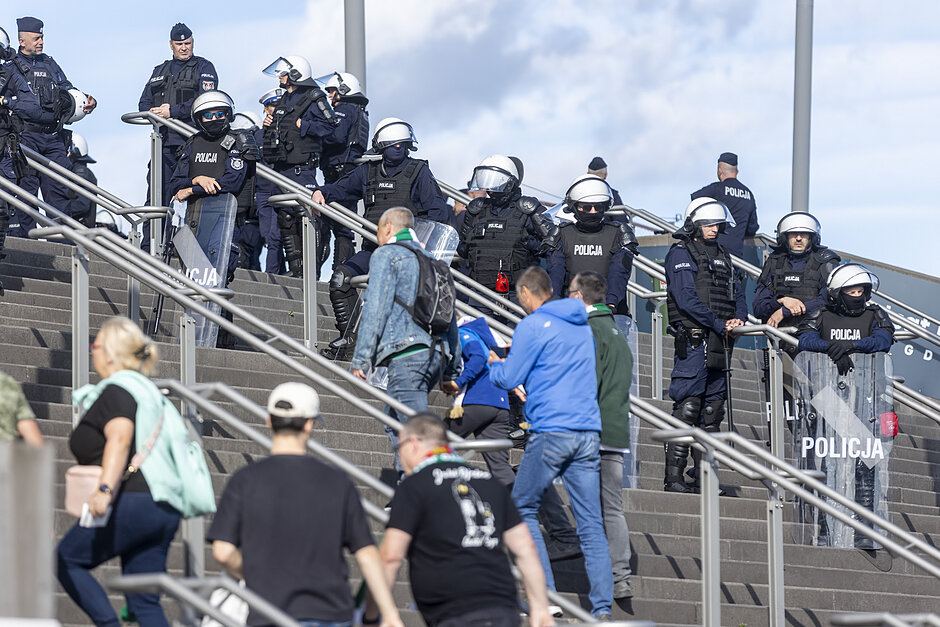 Policjanci w pełnym rynsztunku stoją na schodach, obok nich przechodzą kibice
