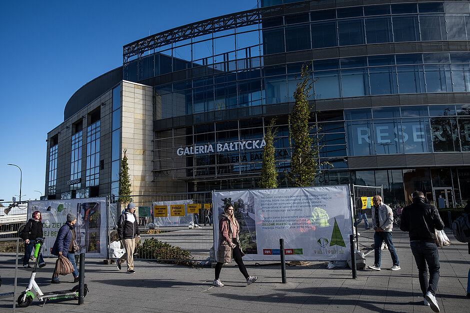 Na placu przed galeria handlową stoi ogrodzenie, za którym ludzie sadza drzewa.