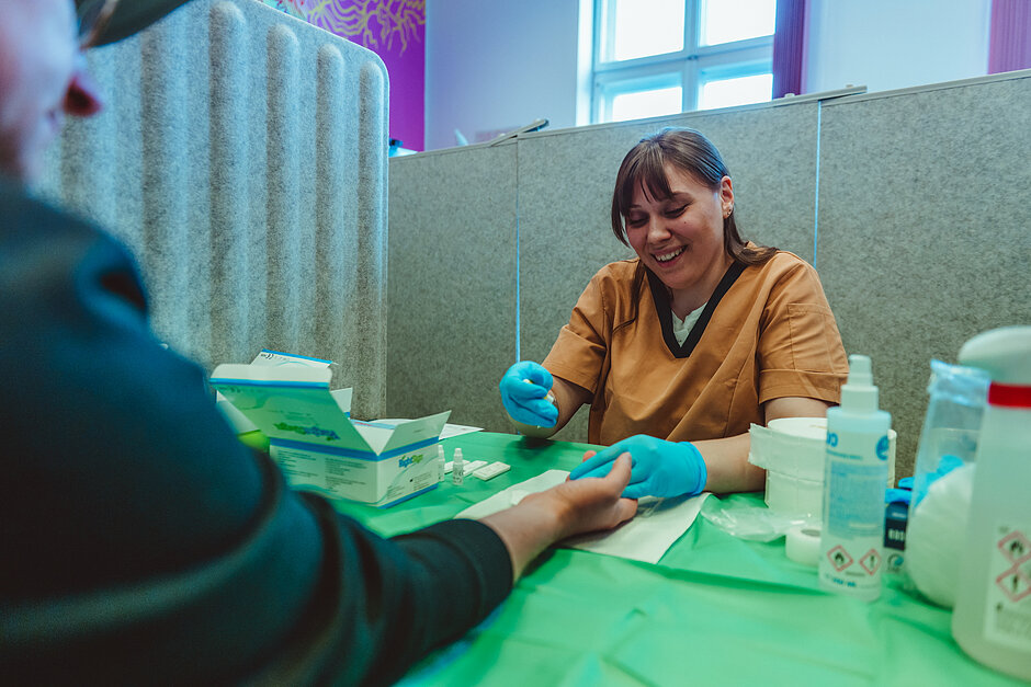 Pracownik medyczny w jednorazowych rękawiczkach wykonuje badanie z kropli krwi u osoby siedzącej przy stole. Na blacie znajdują się testy diagnostyczne, środki dezynfekujące i materiały medyczne. Zdjęcie wykonano w jasnym pomieszczeniu podzielonym przegrodami zapewniającymi prywatność uczestników badań.