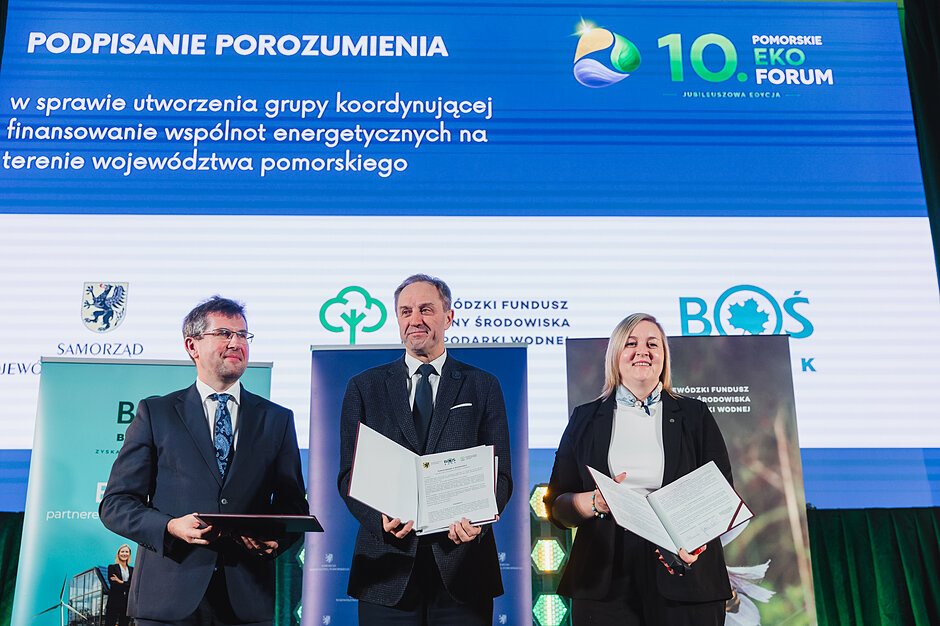 Kobieta i dwóch mężczyzn pozują do zdjęcia z podpisanym porozumieniem na tle ekranu.