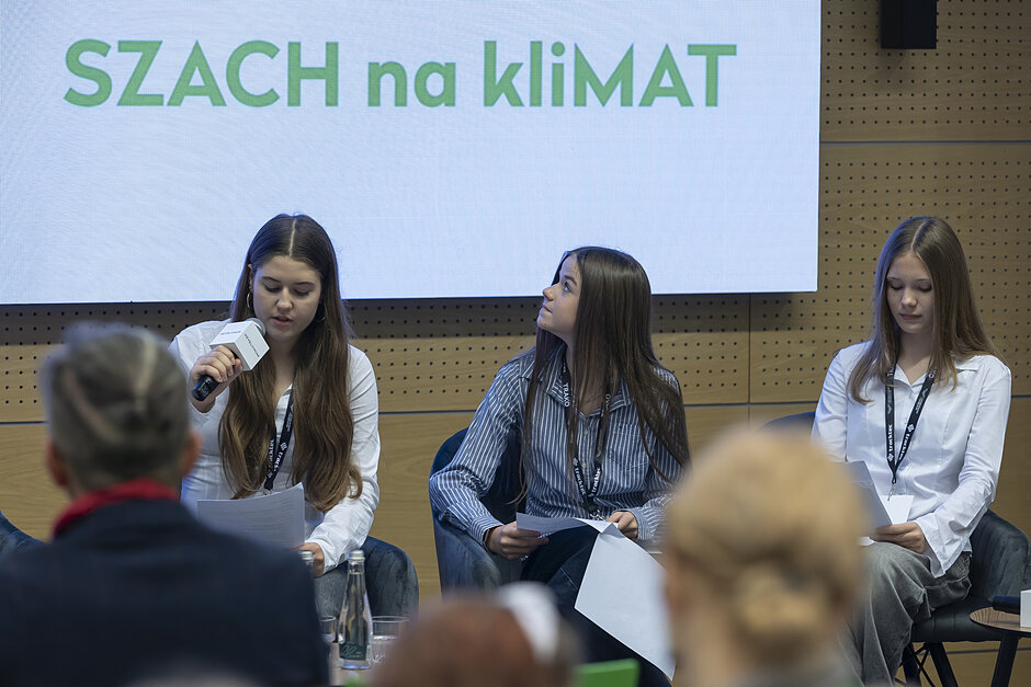 trzy nastolatki siedzą pod ekranem prezentacyjnym na którym napis SZACH na kliMAT, jedna z nich mówi do mikrofonu