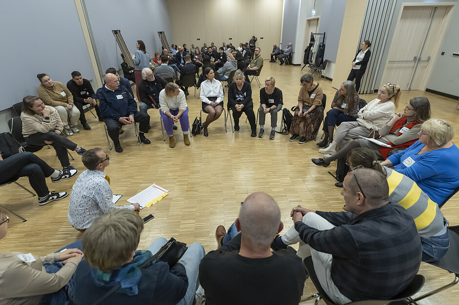 grupa ludzi siedzący w kole, kilkanaście osób. W tle kolejna taka grupa