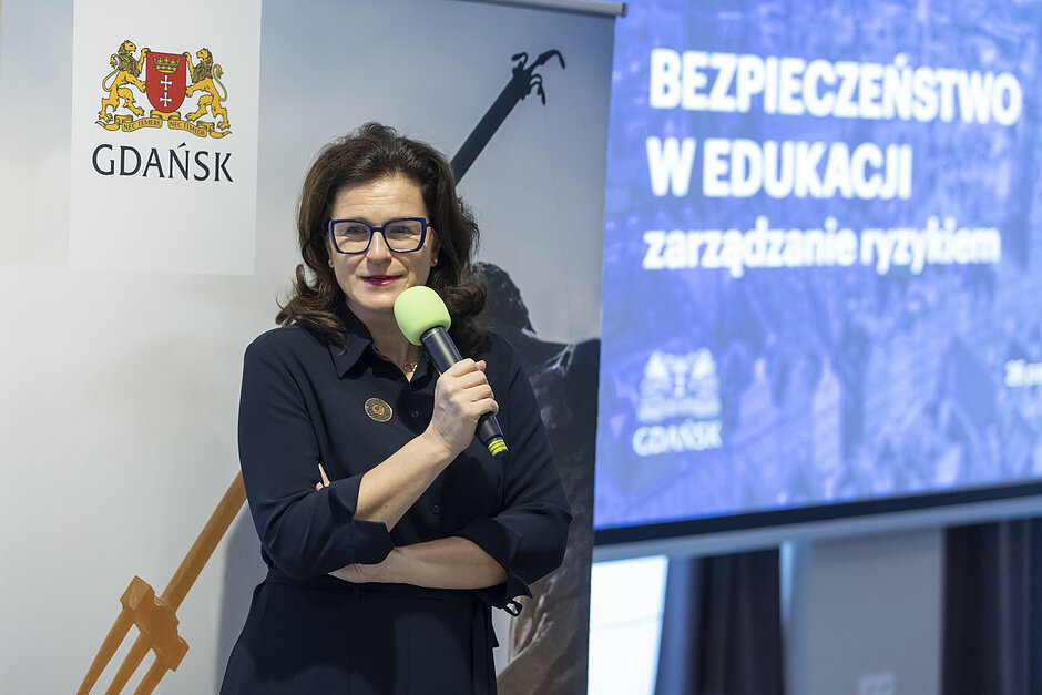 Kobieta z ciemnymi włosami przemawiająca do mikrofonu, za nią plansza z napisem BEZPIECZEŃSTWO W EDUKACJI - zarządzanie ryzykiem