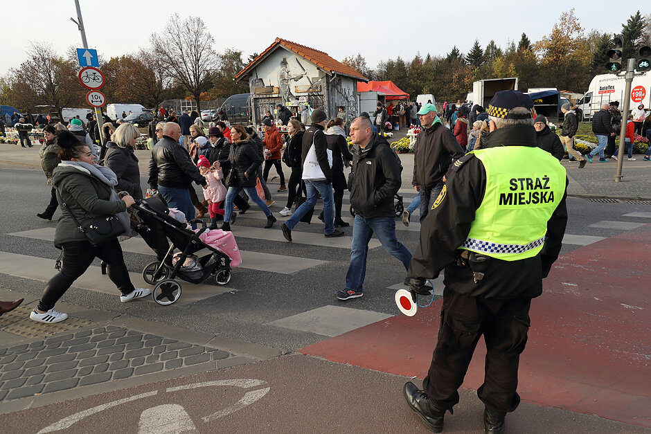 Ludzie przechodzą przez jezdnię przy cmentarzu, a nadzoruje to strażnik miejski.