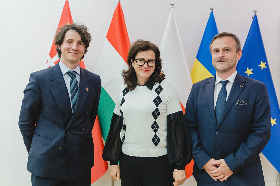 Trzy osoby pozują do zdjęcia na tle flag narodowych, flagi Gdańska i flagi UE. Prezydent Gdańska stoi pośrodku