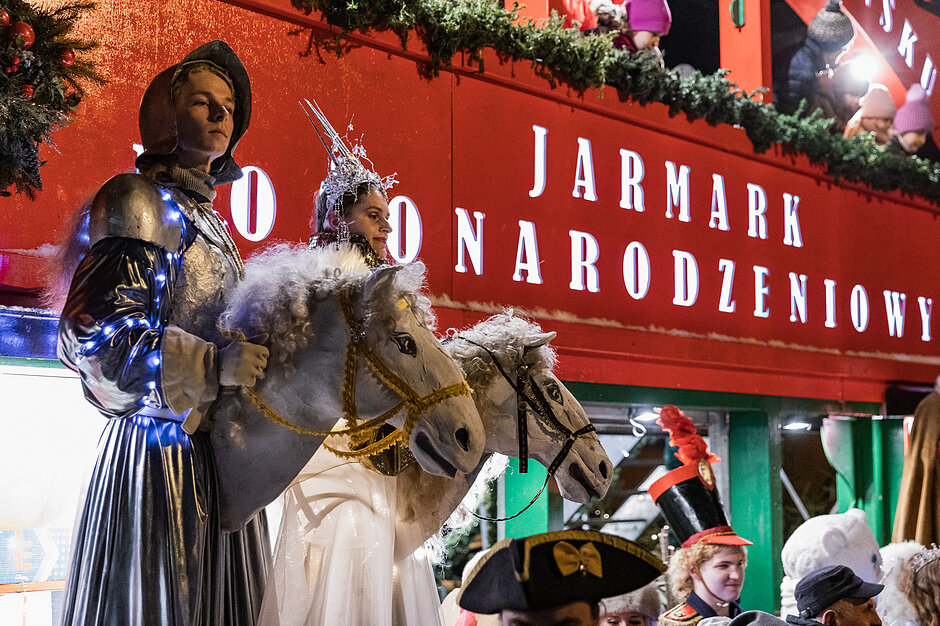 na zdjęciu przebierańcy z drewnianymi łbami końskimi przed sobą, w tle czerwona plansza z napisem jarmark bożonarodzeniowy