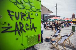 Po lewej stronie zdjęcia fragment zielonej przyczepy z napisem: FURA ŻARCIA. Po prawej dwa leżaki a w tle stoliki i parasole restauracyjne, za nimi budynek