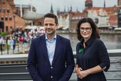 Dwie uśmiechnięte osoby - kobieta i mężczyzna - stoją obok siebie. W tle znajduje się rzeka Motława i panorama Głównego Miasta w Gdańsku, z górującą wieżą Bazyliki Mariackiej