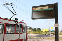 na zdjęciu na pierwszym planie elektroniczna tablica z informacją pasażerską, na której widać numery tramwajów i kierunek odjazdu, w tle widać biało-czerwony tramwaj miejski
