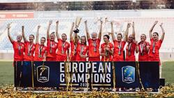 Kilkanaście kobiet w czerwonych strojach sportowych stoi obok siebie, uśmiechają się i machają rękoma, na szyjach mają zawieszone medale. Przed nimi ustawiony jest szeroki baner z napisem European Champions. 