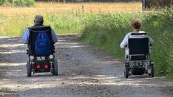 Dwie starsze osoby jadą na wózkach inwalidzkich polną drogą (plecami do obiektywu)