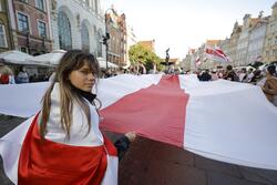 Z lewej stronie zdjęcia młoda dziewczyna odwraca się do aparatu. W tle widoczna wielka flaga Białorusi podtrzymywana przez klika osób. Na jej końcu widoczna Fontanna Neptuna