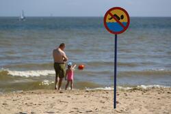 na zdjęciu dorosły mężczyzna i mała dziewczynka w strojach kąpielowych, idą brzegiem morza, po piasku, na pierwszym planie znak informujący o zakazie pływania