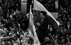Czarno-białe zdjęcie: tłum ludzi, w środku mężczyzna z wąsami (bokiem), w prawej dłoni trzyma flagę narodową, w lewej trzyma mikrofon
