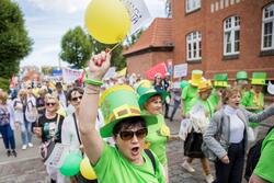 na pierwszym planie widać kobitę z balonikiem w ręku, ma na głowie papierowy zielony kapelusz, ma też na sobie zieloną koszulkę z krótkim rękawem, w tle tłum osób, głównie seniorów, wszyscy są barwnie ubrani, idą w paradzie