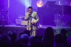 czarnoskóry mężczyzna w garniturze mówi do mikrofonu na scenie, fioletowe światło