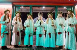 grupa ubranych na biało ośmiu dziewcząt śpiewa na placu nocą, na głowach mają wianki, a niektóre korony ze świeczek