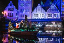 Zdjęcie zrobione po zmroku. Motławą płynie łódź ze św. Mikołajem na pokładzie. Na ściany nadbrzeżnych domów pada fioletowe światło, wokół jest mnóstwo iluminacji