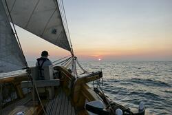 żaglowiec płynie po morzu przy zachodzie słońca, dziób i żagiel, jedna osoba na pokładzie