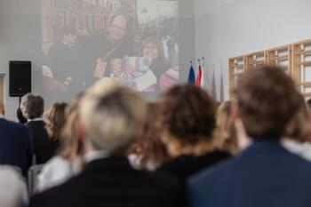 z przodu kilka rzędów ludzi siedzących na krzesłach, na ścianie ekran ze zdjęciem Pawła Adamowicza