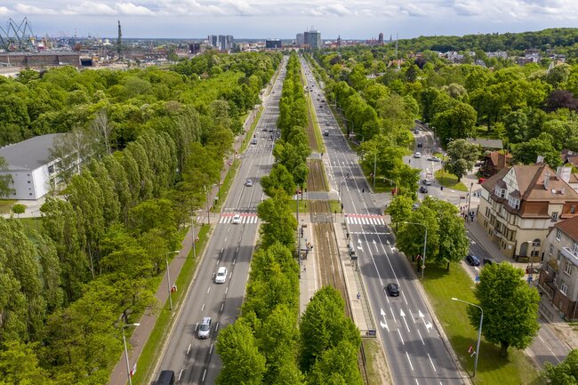 zdjęcie z drona, widać główną trasę samochodową przez miasto, jezdnie mają po trzy pasy, w każdym kierunku rozdzielone są pasmami zielonych drzew, w tle widać kamienice