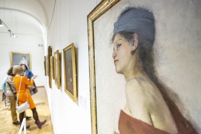 duży portret kobiety w złotych ramach na ścianie w galerii, w tle mniejsze obrazy i oglądający