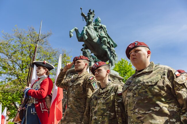 na zdjęciu trzech młodych żołnierzy w mundurach i jedne mężczyzna w historycznym stroju wojskowym, nad nimi w tle stoi pomnik króla siedzącego na koniu