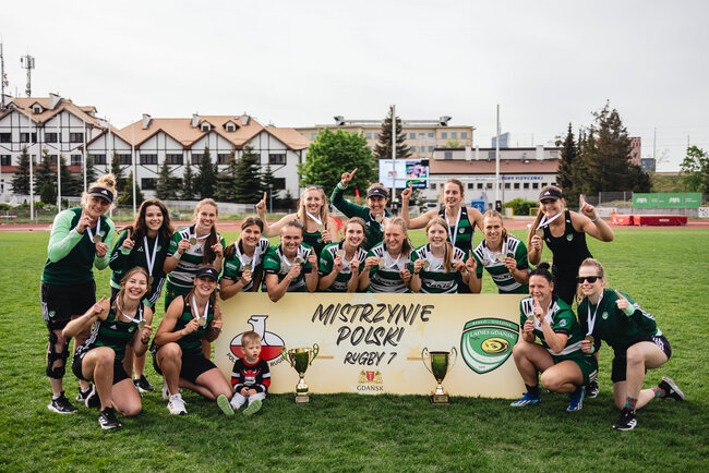 Grupa zawodniczek rugby pozuje do zdjęcia po zwycięskim meczu, który zapewnił im mistrzostwo Polski. Kobiety są uśmiechnięte i pokazują palcem numer 1. Między nimi stoi baner z napisem Mistrzynie Polski rugby 7 oraz złoty puchar.