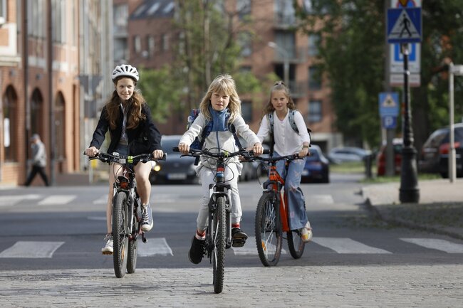 Na zdjęciu widać trzy młode dziewczyny, które jadą na rowerach ulicą w mieście. Wszystkie mają na sobie kaski i plecaki, co sugeruje, że mogą być w drodze do szkoły lub z niej. Jazda odbywa się w pobliżu przejścia dla pieszych, a w tle widać ceglany budynek oraz kilka zaparkowanych samochodów. Dziewczyny wyglądają na skoncentrowane i radosne z jazdy na rowerach. 