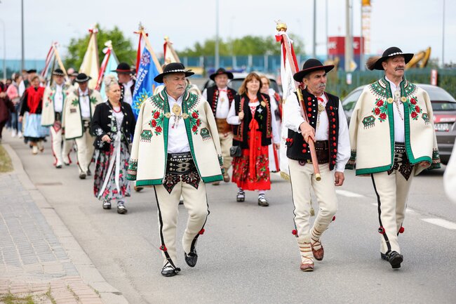 Na zdjęciu widać grupę osób maszerujących w tradycyjnych, regionalnych strojach, prawdopodobnie podczas jakiegoś festiwalu lub parady. Osoby te noszą bogato zdobione, kolorowe ubrania i szarfy, a mężczyźni trzymają dekoracyjne flagi oraz noszą kapelusze z piórami