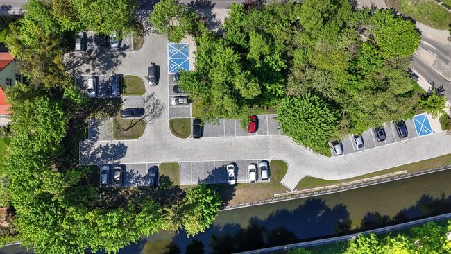 zdjęcie z drona, widać parking z wyznaczonymi miejscami postojowymi, kilka stojących samochodów, a wokół zielone drzewa i trawniki
