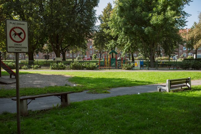 fragment parku, widać alejkę spacerową z asfaltu, przy niej stoi ławka, w tle dużo zielonych drzew i plac zabaw, po lewej widać znak o zakazie wyprowadzania psów