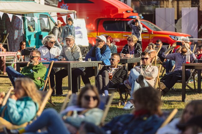 Zdjęcie przedstawia grupę ludzi siedzących na zewnątrz podczas jakiegoś wydarzenia, najprawdopodobniej festiwalu lub targów. W tle widać czerwony pojazd strażacki, a uczestnicy siedzą przy stolikach oraz na leżakach, niektórzy z nich patrzą na coś przed sobą lub rozmawiają ze sobą.