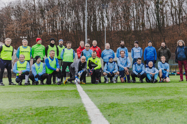 Na zdjęciu znajduje się grupa osób na boisku piłkarskim, podzielonych na dwie drużyny - jedna w niebieskich strojach, a druga w zielonych kamizelkach treningowych. W tle widoczny jest las, a uczestnicy wyglądają na zadowolonych, gotowych do wspólnej gry lub właśnie po zakończonym meczu.