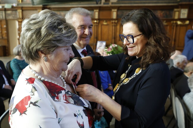 Na zdjęciu uśmiechnięta kobieta w ciemnym stroju z ozdobnym naszyjnikiem przypina odznaczenie starszej kobiecie w eleganckiej, kwiecistej sukni, która nosi perłowy naszyjnik. W tle widać mężczyznę w garniturze i innych uczestników uroczystości w bogato zdobionym wnętrzu
