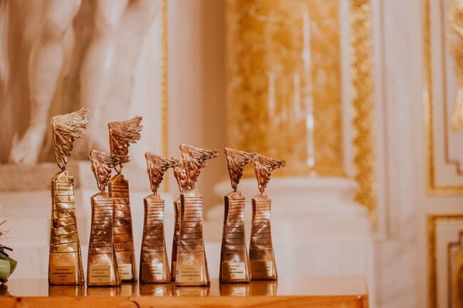 Na zdjęciu widać kilka statuetek z charakterystycznymi skrzydłami ustawionych na drewnianym blacie. W tle znajduje się eleganckie wnętrze z pozłacanymi zdobieniami i klasycznymi detalami architektonicznymi. 