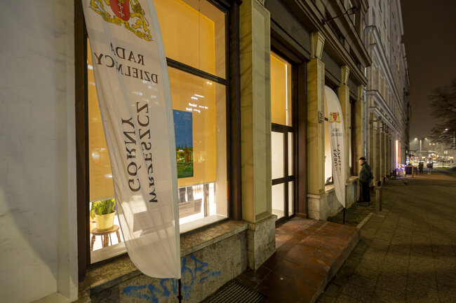 Wieczorne zdjęcie przy latarniach miejskich budynku przed którym powiewa baner Rady Dzielnicy.