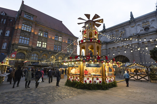 tętniący życiem jarmark bożonarodzeniowy, znajduje się przed dużym budynkiem z czerwonym dachem i ozdobnymi dekoracjami. W centrum znajdują się małe drewniane stragany zdobione czerwonymi i białymi kwiatami i konstrukcją przypominającą wiatrak na górze.