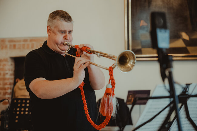 Mężczyzna gra na trąbce we wnętrzu sali, trzymając instrument ozdobiony czerwonym sznurem i frędzlami. W tle widoczne są pulpity z nutami oraz fragmenty wyposażenia wnętrza, co sugeruje próbę lub występ muzyczny. 