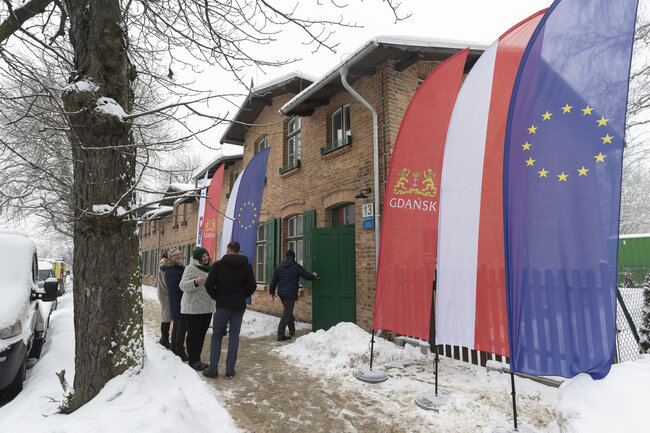 Ceglany dom w śnieżnej scenerii. Przed domem flagi Gdańska, Polski i Unii Europejskiej oraz kilka osób.