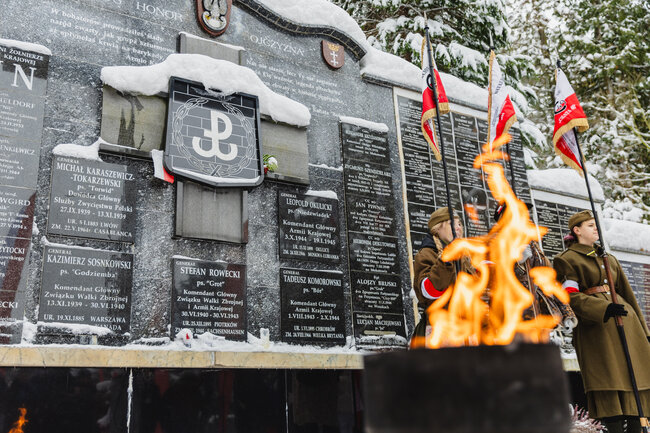 Na zdjęciu widać uroczyste obchody Narodowego Dnia Pamięci Żołnierzy Armii Krajowej, odbywające się przy Kwaterze Akowskiej na Cmentarzu Łostowickim w Gdańsku. Przed pamiątkową ścianą z nazwiskami dowódców płonie symboliczny znicz, a obok czuwają rekonstruktorzy w historycznych mundurach z polskimi flagami.