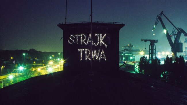 Nocne zdjęcie terenu stoczni zrobione z poziomu dachów hal produkcyjnych. Na pierwszym planie na dachu wielki napis białą farbą Strajk Trwa. W tle panorama miasta i dźwigi stoczniowe