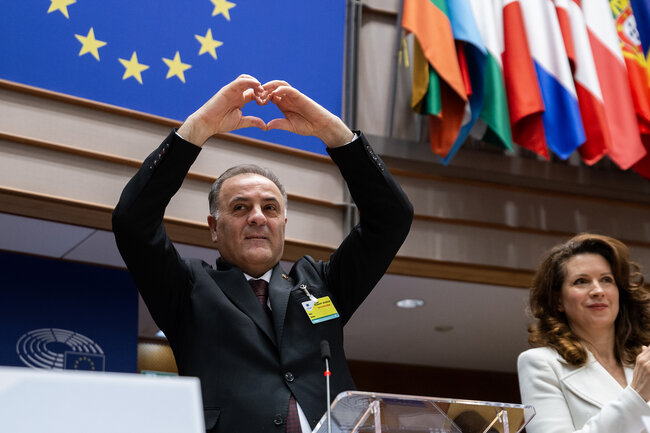 Mężczyzna na mównicy w sali obrad stoi i pokazuje palcami serce.