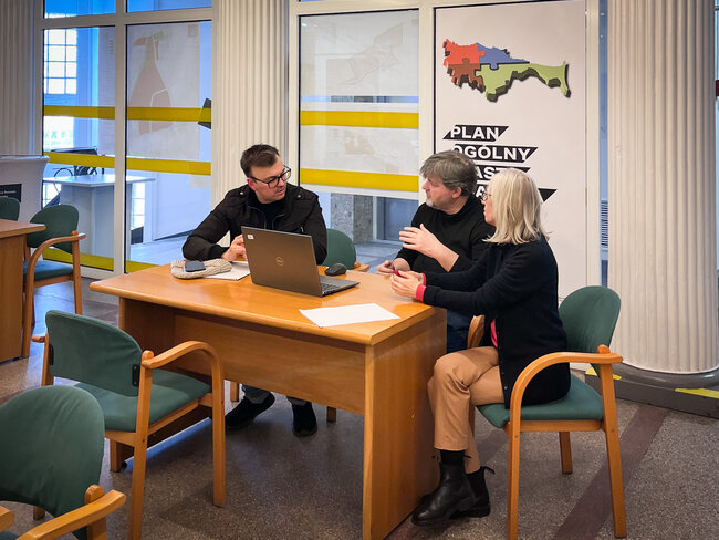 trzy osoby ubrane na czarno siedzą przy brązowym biurku i rozmawiają